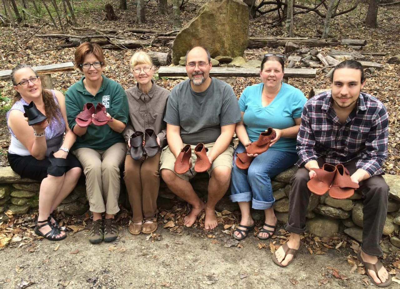 Small group of students sitting on a stone wall at Earthaven Ecovillage with the shoes they made in a shoemaking workshop